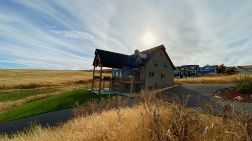 A modern house on a hillside with a sunlit sky, surrounded by dry grass and distant homes.